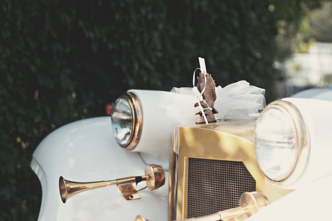 Classic white car front with a gold grille, brass trumpet horn, and an eagle figurine draped in white ribbons.