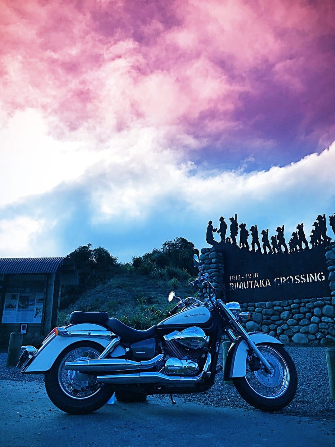 Chrome motorcycle parked by the Rimutaka Crossing memorial under a purple and pink sunset sky with silhouettes.