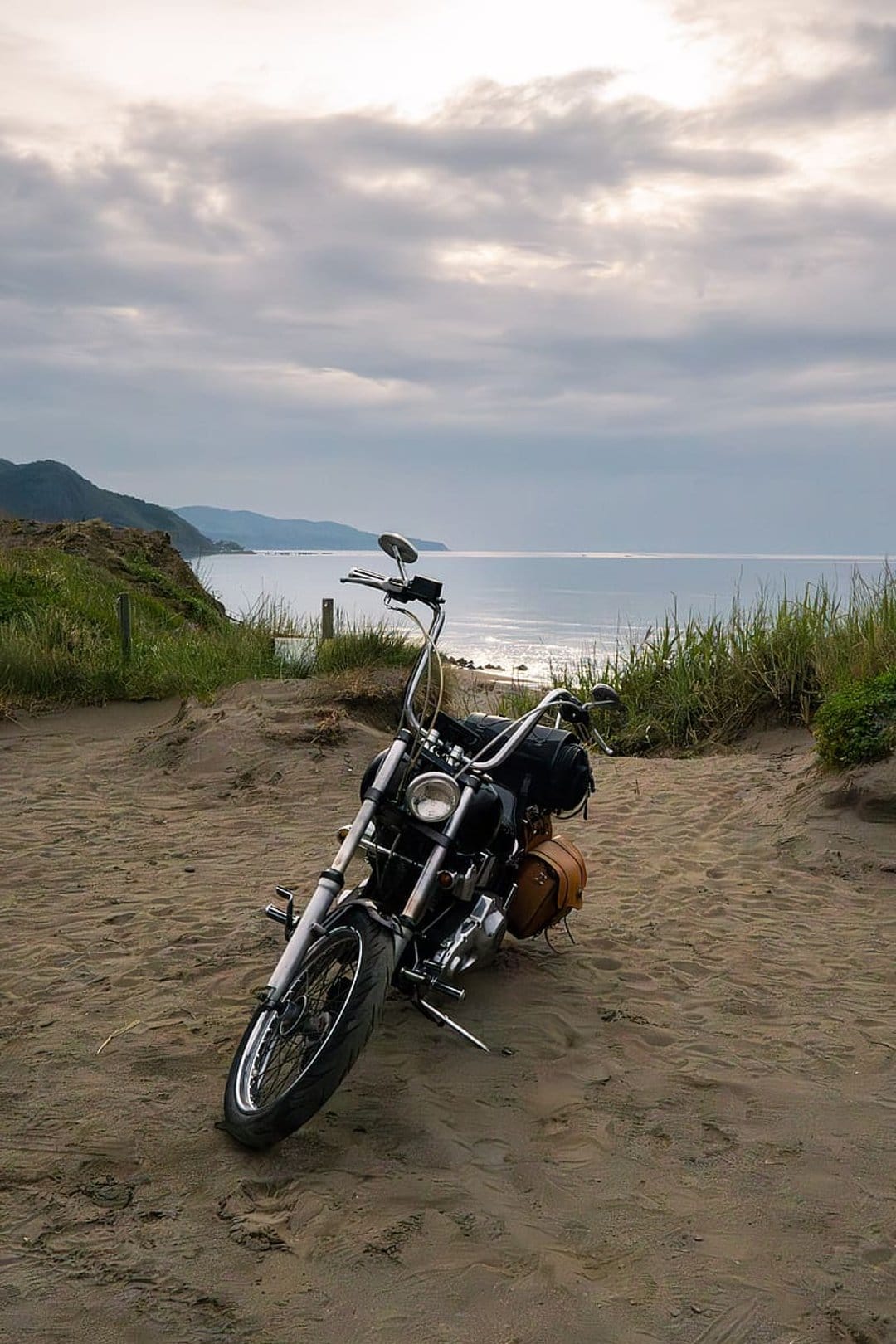 Black vintage motorcycle with chrome accents parked on sandy dunes beside beach grass and a calm coastal ocean.