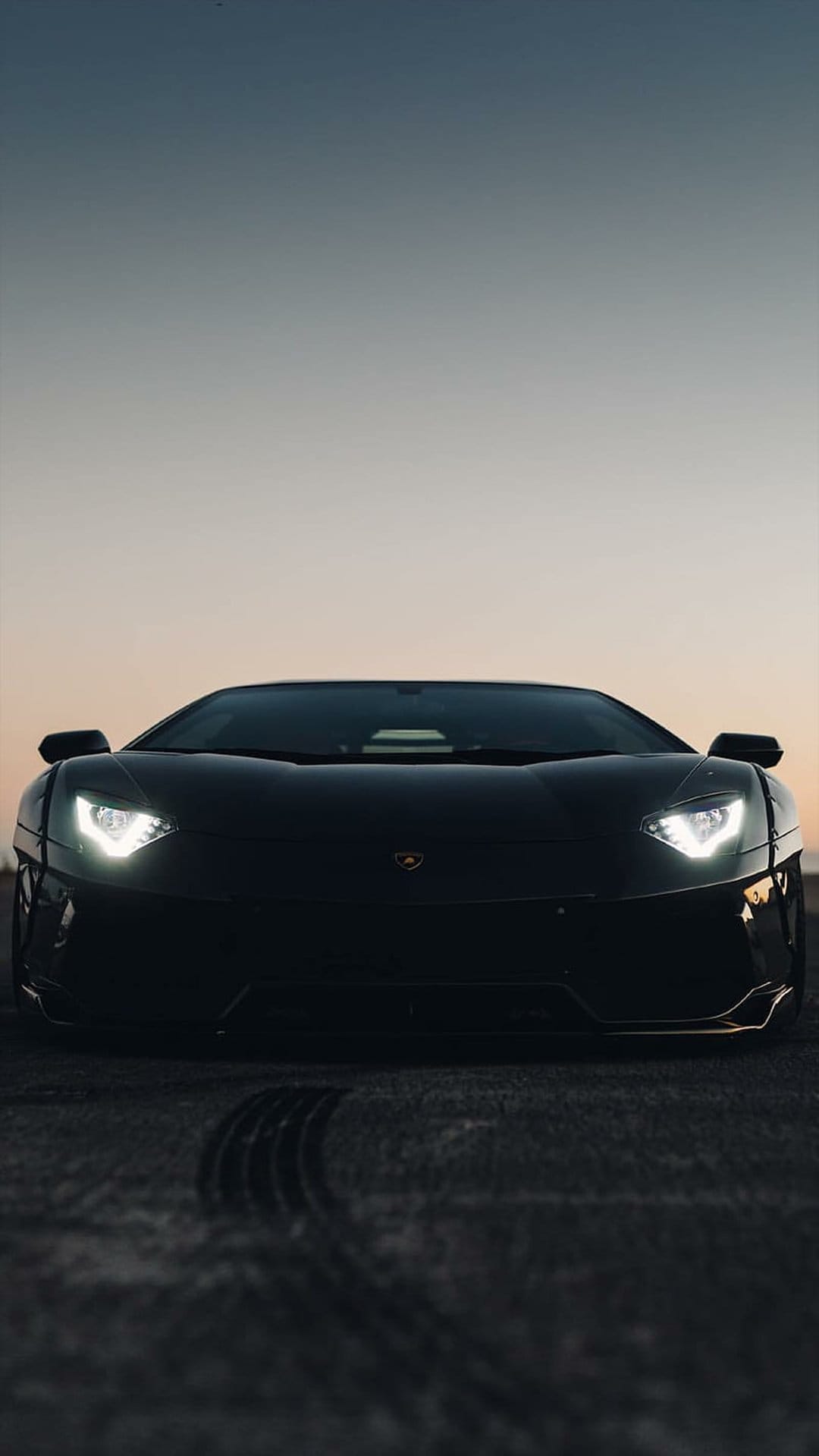 Black Lamborghini Aventador with glowing LED headlights on dark asphalt under a moody twilight sky.