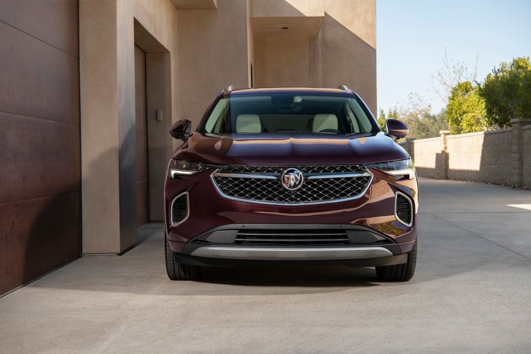 Maroon Buick Envision SUV front view in a modern driveway with sunlight hitting the chrome grille.