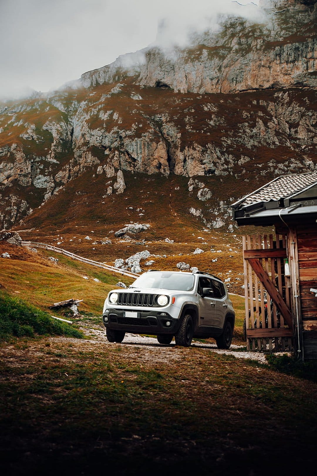 Silver Jeep Renegade on a dirt path next to a rustic wooden cabin under a cloudy, mountainous alpine sky.