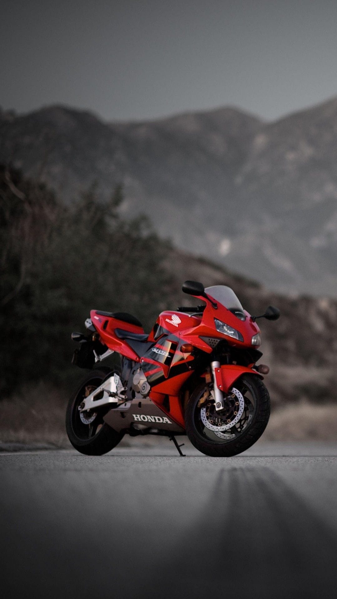 Red Honda CBR sportbike parked on an asphalt mountain road with foggy hills and green trees in the background.