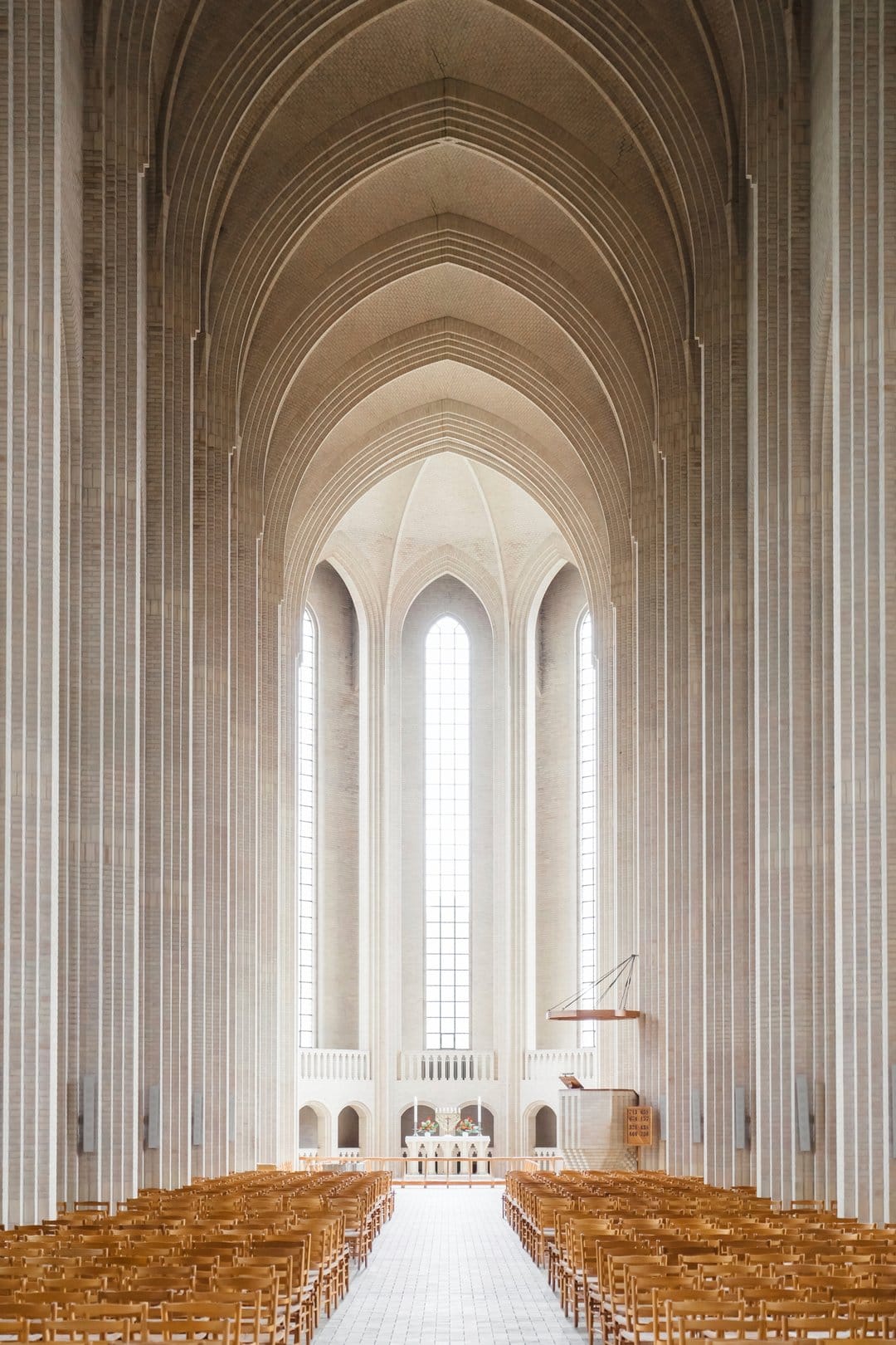 High vaulted ceilings and pointed concrete arches of a minimalist church interior with wooden pews and soft light.