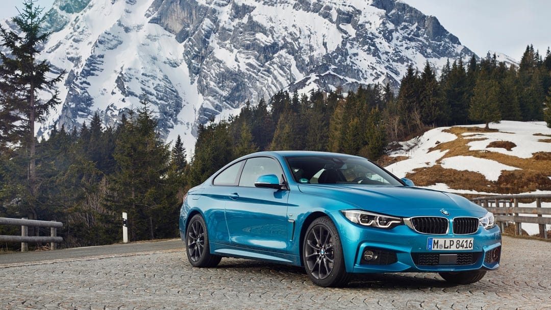 Blue BMW coupe on a cobblestone road with a backdrop of snow-covered mountains and evergreen trees.