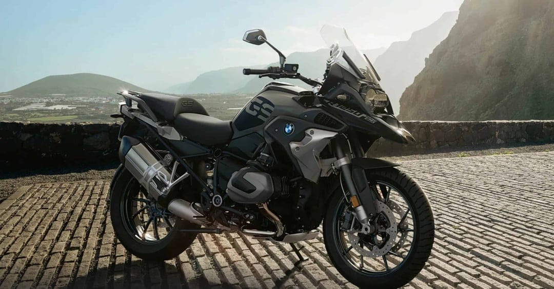 BMW R 1250 GS motorcycle on a cobblestone mountain road with sunlight and distant peaks in the background.
