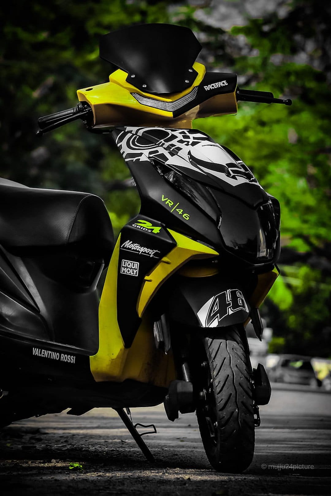 Yellow VR46 scooter with number 46 racing decals parked against a blurred green garden landscape backdrop.