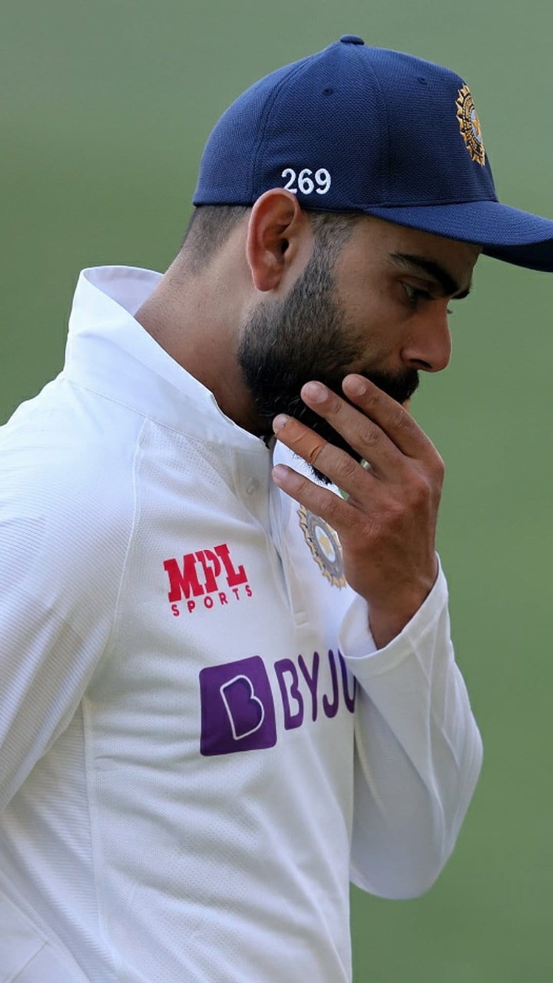 Virat Kohli wearing a white India Test jersey and blue cap with a hand near his chin in a thoughtful pose.