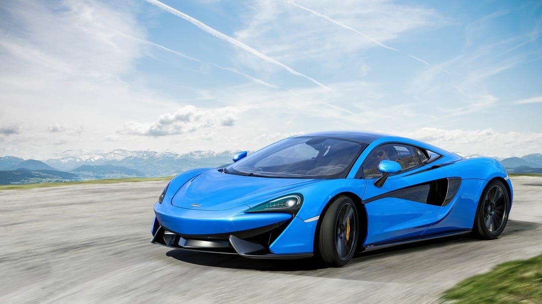 Blue McLaren supercar driving fast on a scenic mountain road with snow-capped peaks and a bright sky.