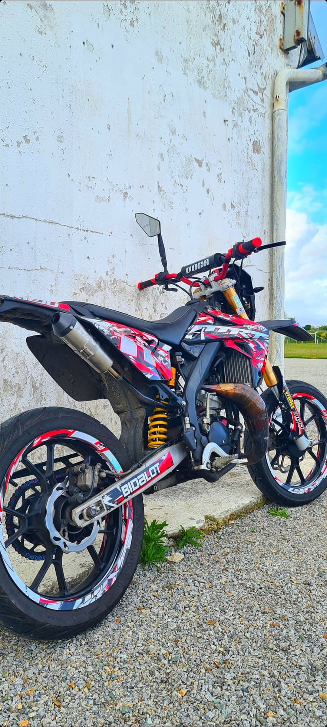 Custom red and black dirt bike with yellow shock absorber parked in front of a weathered white outdoor wall.