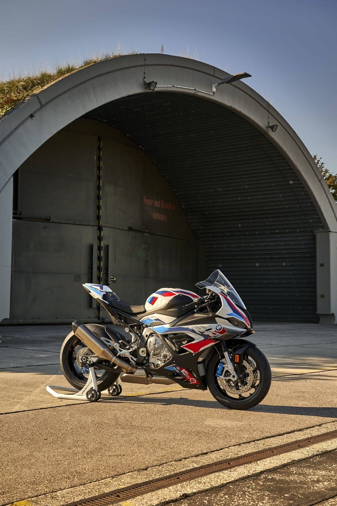 BMW M 1000 RR motorcycle parked in front of a large concrete aircraft shelter during golden hour sunlight.