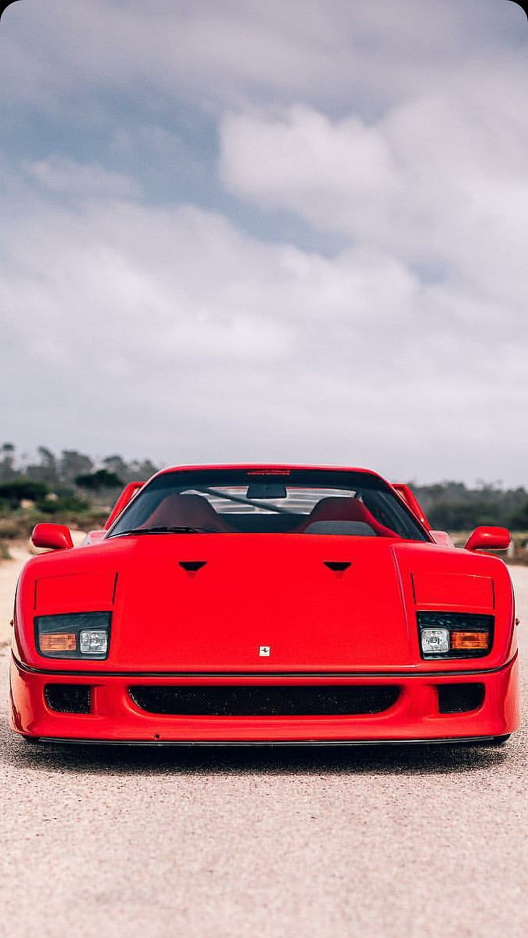 Ferrari F40 front fascia with pop-up headlights parked on a paved road beside green coastal vegetation.