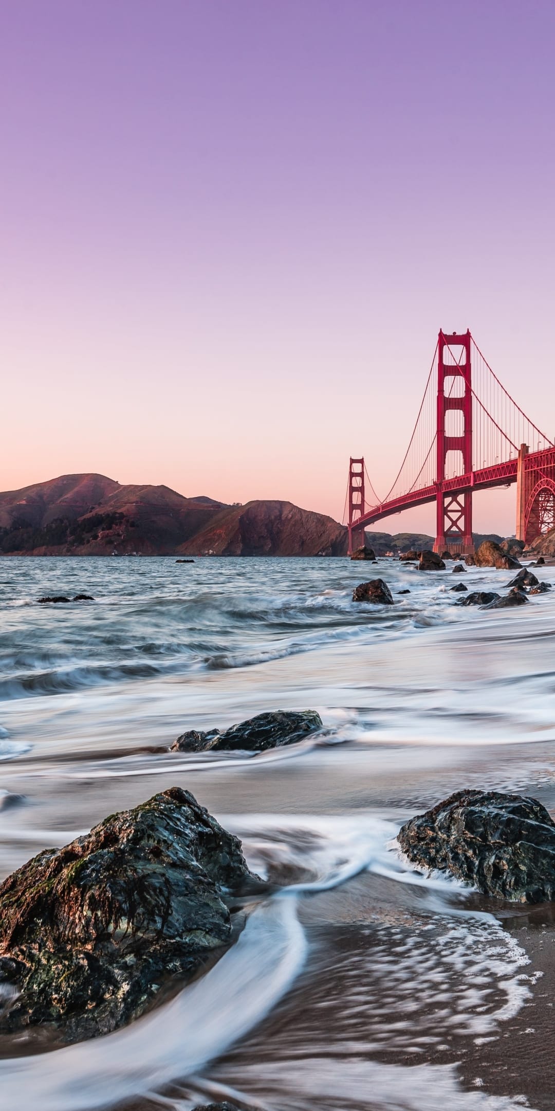 Golden Gate Bridge towers over crashing waves and mossy rocks under a lavender and rose twilight sky.