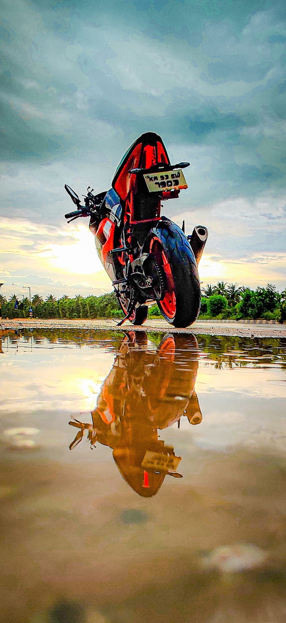 KTM motorcycle performs a wheelie on a wet road with a symmetrical reflection in a puddle under a sunset sky.