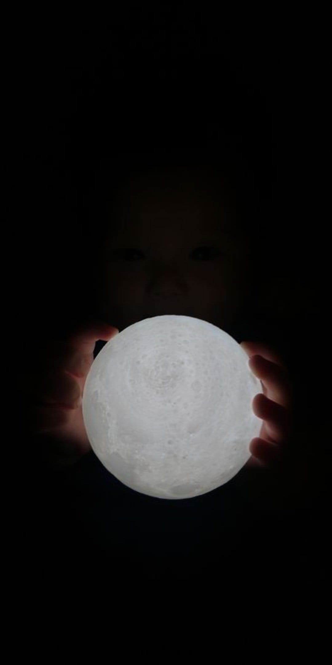 Child's hands hold a glowing moon orb in darkness with subtle eyes visible, providing soft light in a black scene.