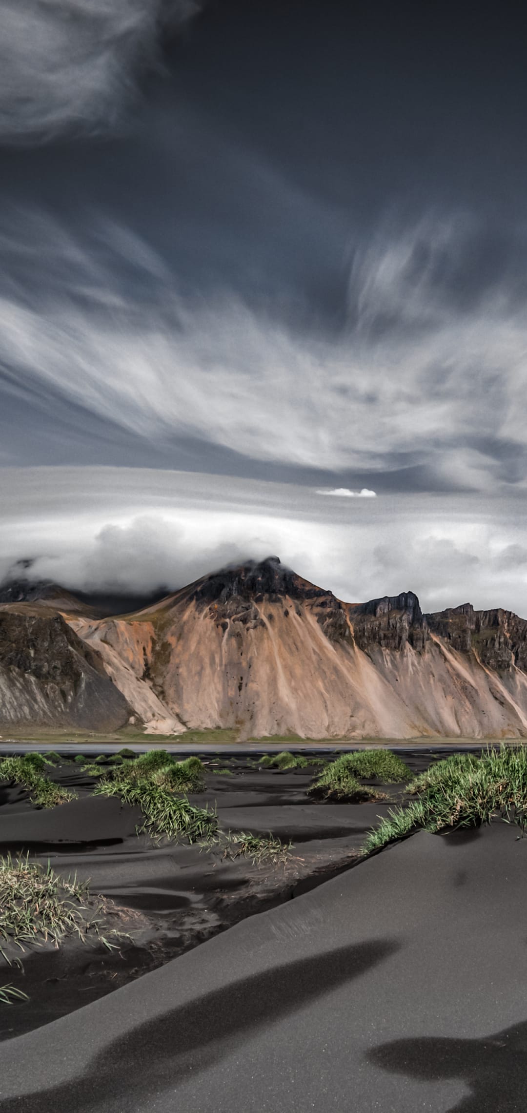 Vestrahorn mountain peaks rise behind black sand dunes and green grass tufts under a swirling, cloudy sky.