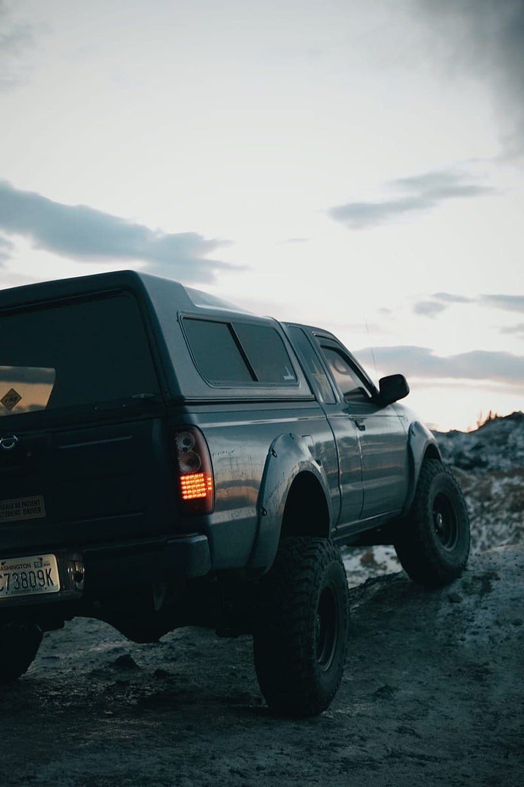 Grey Nissan Frontier pickup truck with a camper shell parked on a dirt path during a moody evening sunset.