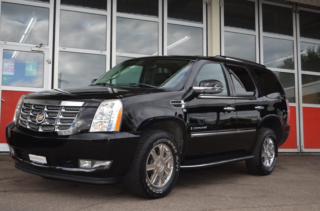 Black Cadillac Escalade with chrome grille and wheels parked in front of a building with glass windows.