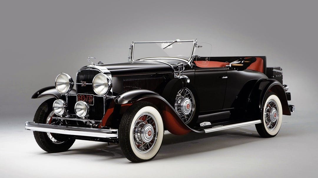 Black vintage convertible car with red interior, chrome grille, and whitewall tires on a grey background.