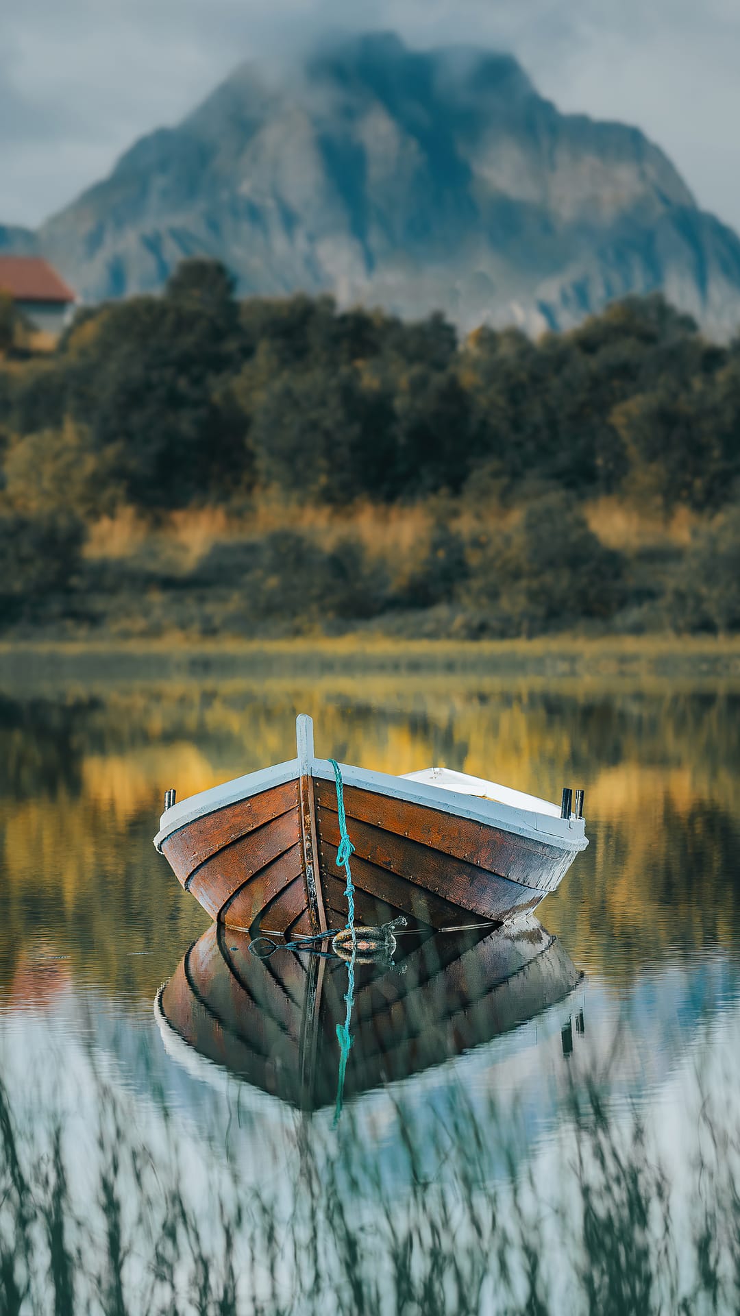 Wooden rowboat on a glass-like lake reflecting a misty mountain and green shoreline under a cloudy sky.