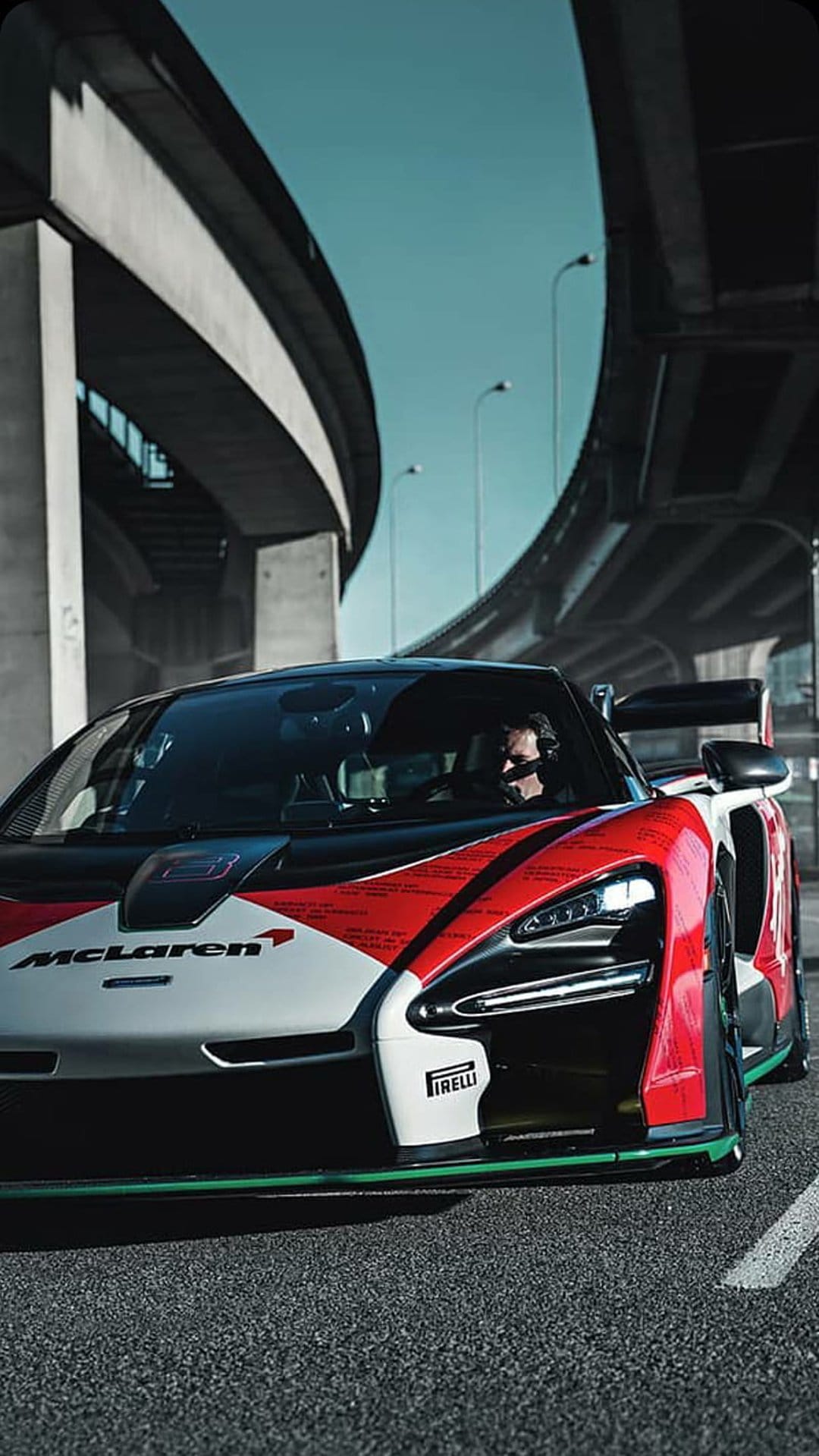 McLaren Senna GT race car with red livery parked under concrete highway overpasses against a blue sky.