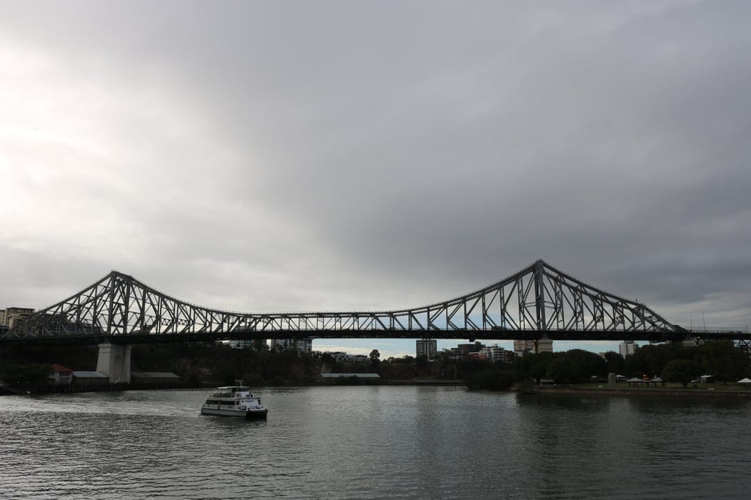 Story Bridge spanning the Brisbane River with a ferry on the water under a dramatic grey cloudy sky.
