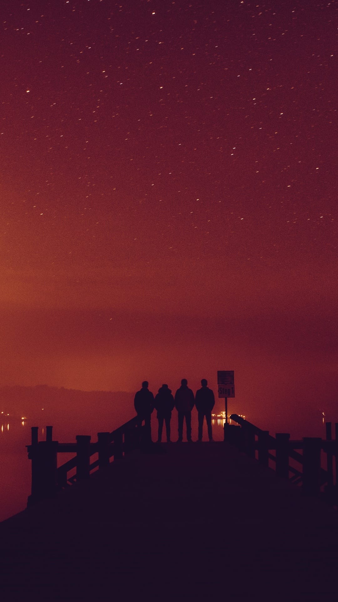 Four people stand in silhouette on a wooden dock under a vibrant red and orange starry night sky over dark water.