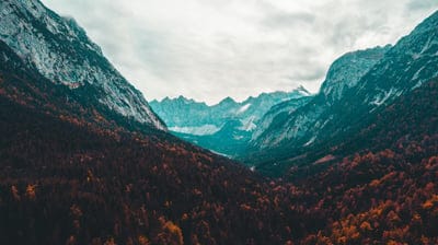 Jagged grey peaks tower over a dense valley of red and orange autumn trees beneath a cloudy, dramatic sky.
