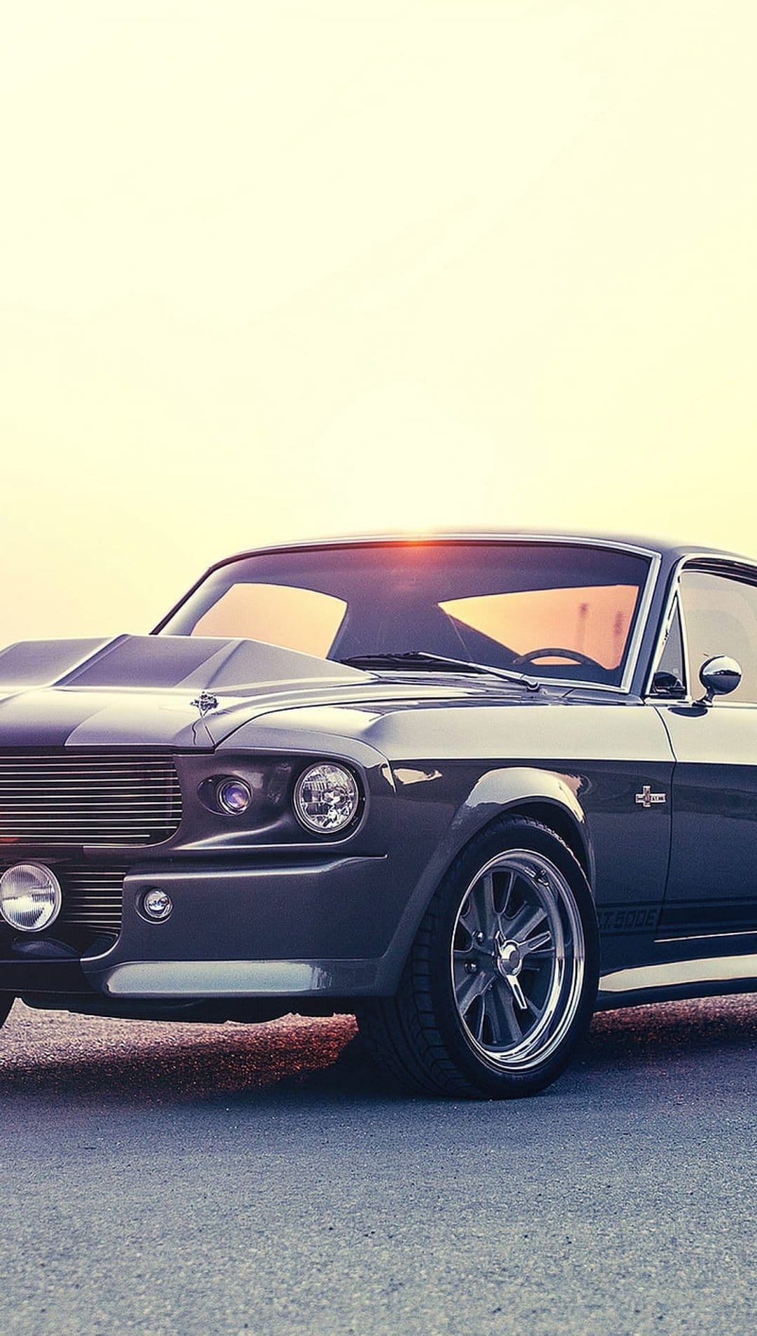 Gray Shelby GT500 Mustang with hood scoop and round headlights parked on asphalt during a warm, golden sunset.