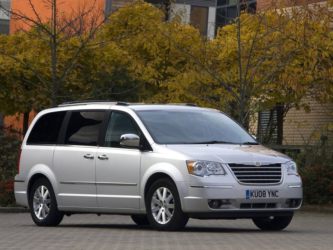 Silver Chrysler Grand Voyager minivan parked on a stone plaza with golden autumn trees in the background.