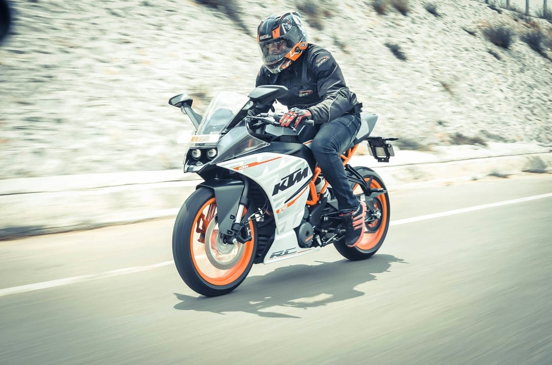Motorcyclist in a full helmet leans on an orange KTM RC sportbike while riding along a blurred hillside road.