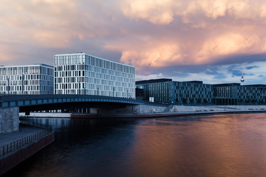 Modern white buildings with geometric facades reflected in a calm river under a pink and purple sunset sky.