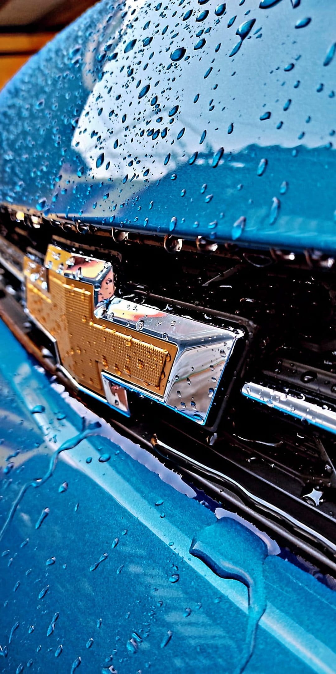 Blue Chevrolet emblem on a wet car grille with clear raindrops and metallic reflections in a vertical macro view.