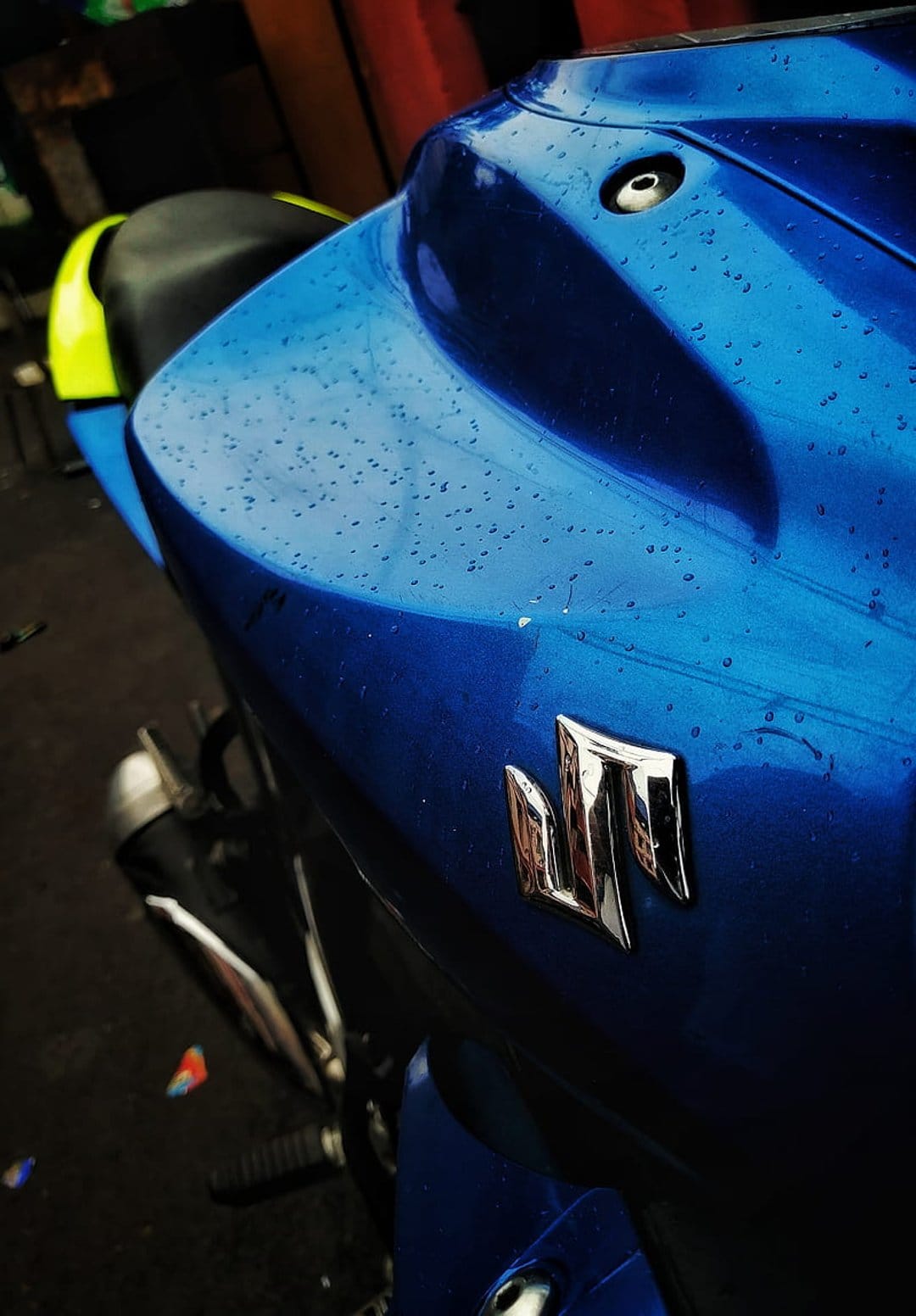 Blue Suzuki motorcycle bodywork covered in water droplets with a silver chrome logo reflecting a rainy exterior.