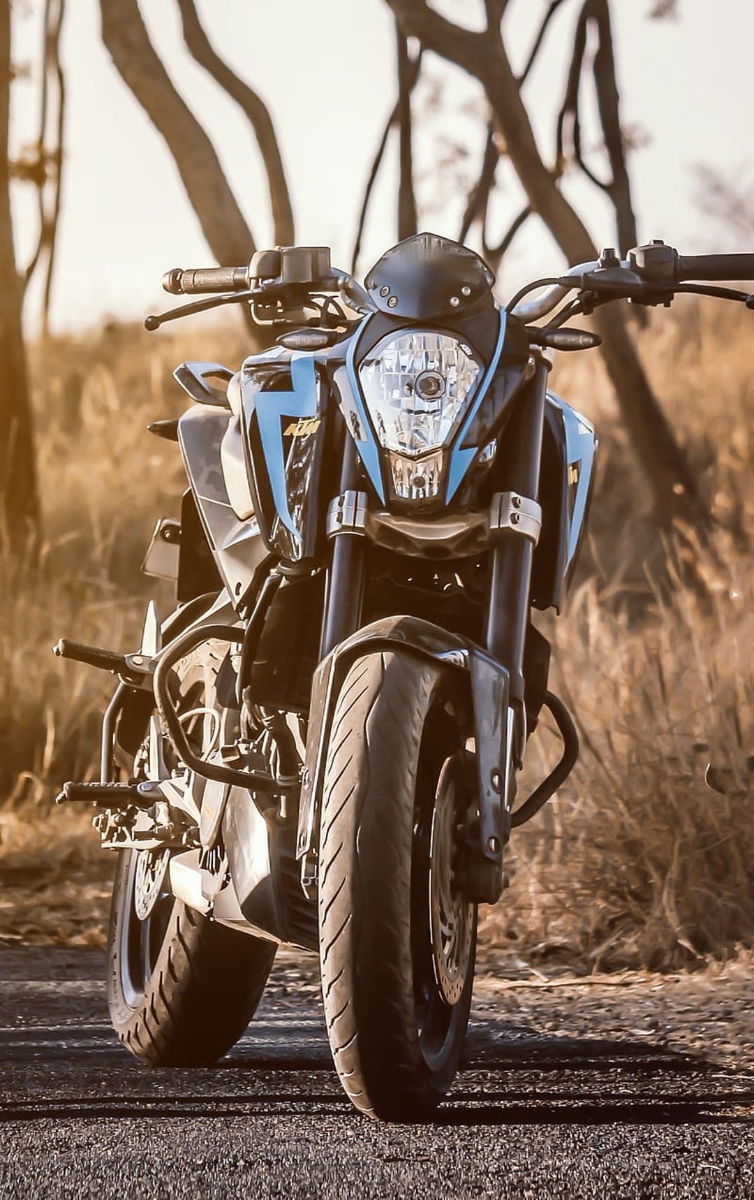 Blue KTM Duke sportbike features close-up front wheel and handlebars against a backdrop of dry grass and trees.