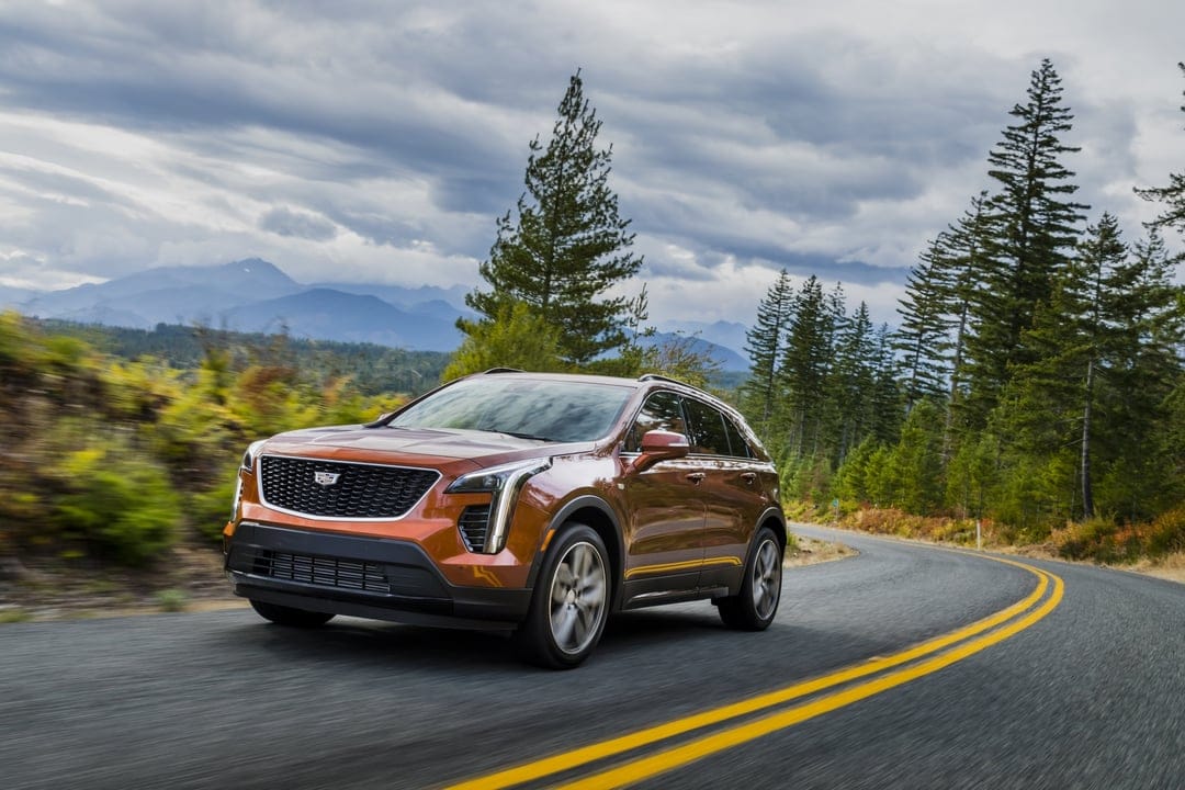 Orange Cadillac SUV driving on a curved forest road with motion blur and distant mountains under cloudy skies.