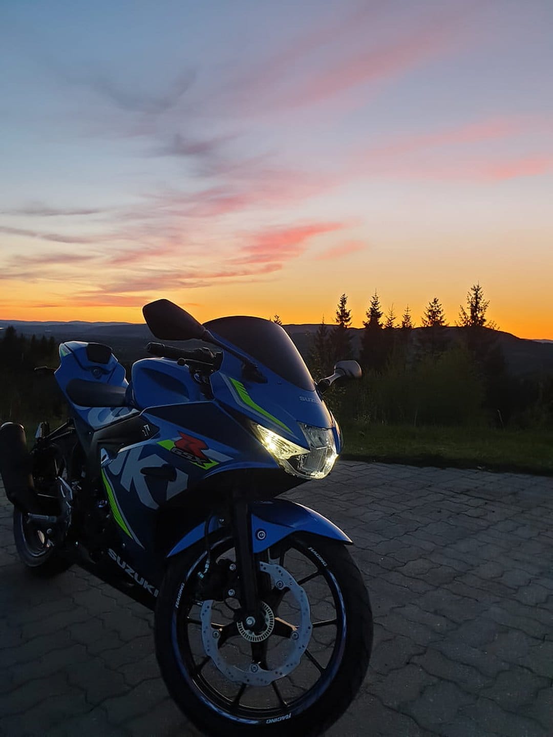 Blue Suzuki motorcycle with lit headlights parked on a road during a vivid orange and pink mountain sunset.