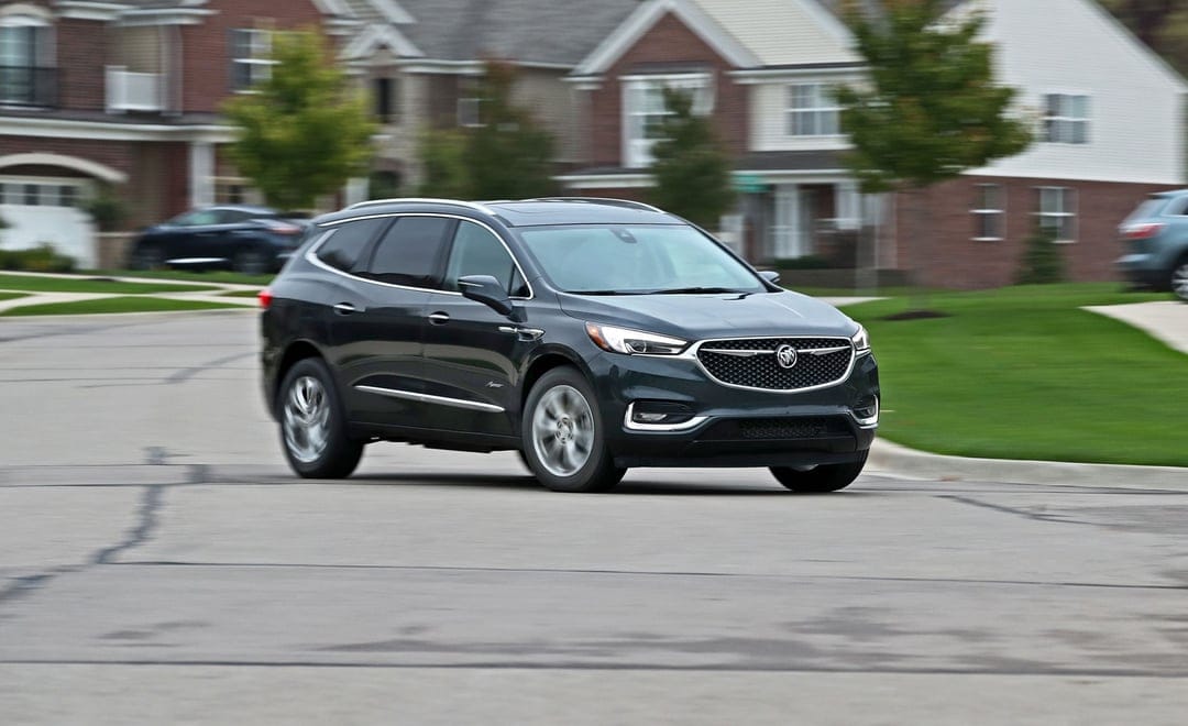Dark grey Buick Enclave SUV in motion on a suburban road with blurred residential houses and green lawns.