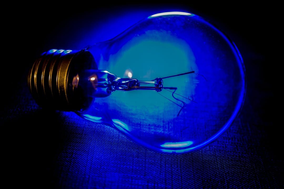 Blue glowing light bulb with a brass base lying horizontally on a black textured surface in low light.