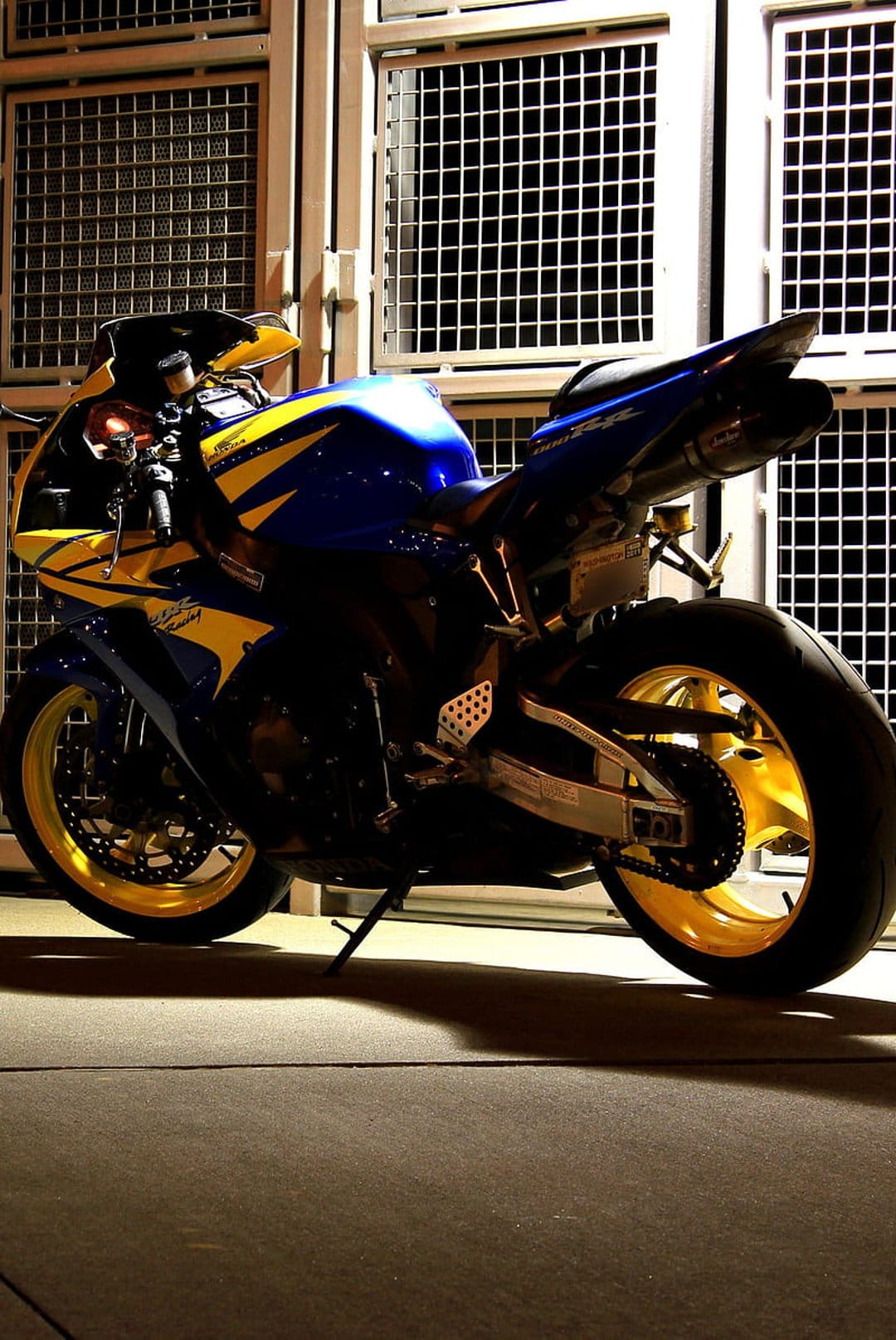 Blue and yellow sportbike with gold wheels parked in front of a gray grid wall with dramatic shadows and lighting.