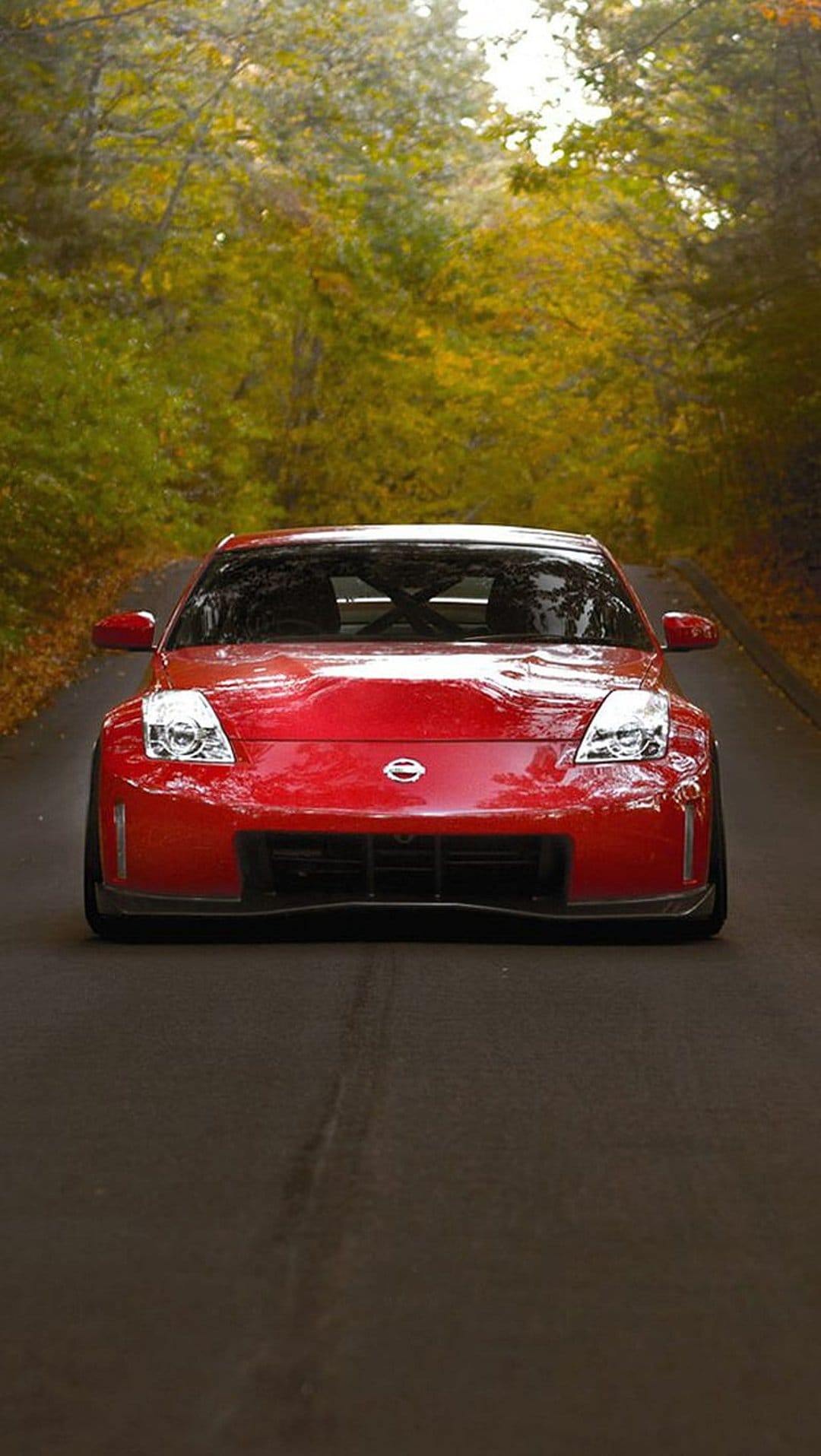 Red Nissan 350Z sports car parked on an asphalt road covered in yellow autumn leaves within a dense forest.