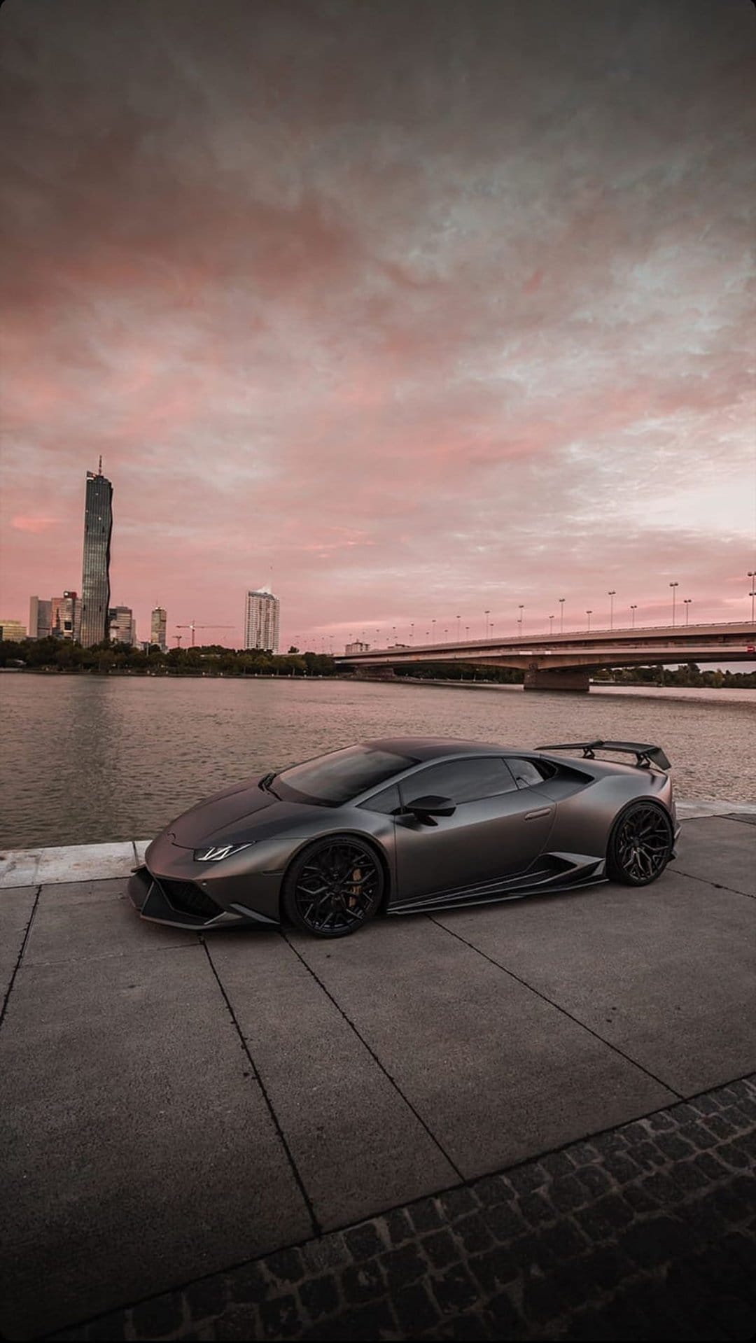 Dark grey Lamborghini on a waterfront promenade under a pink sunset sky with a distant city skyline and bridge.