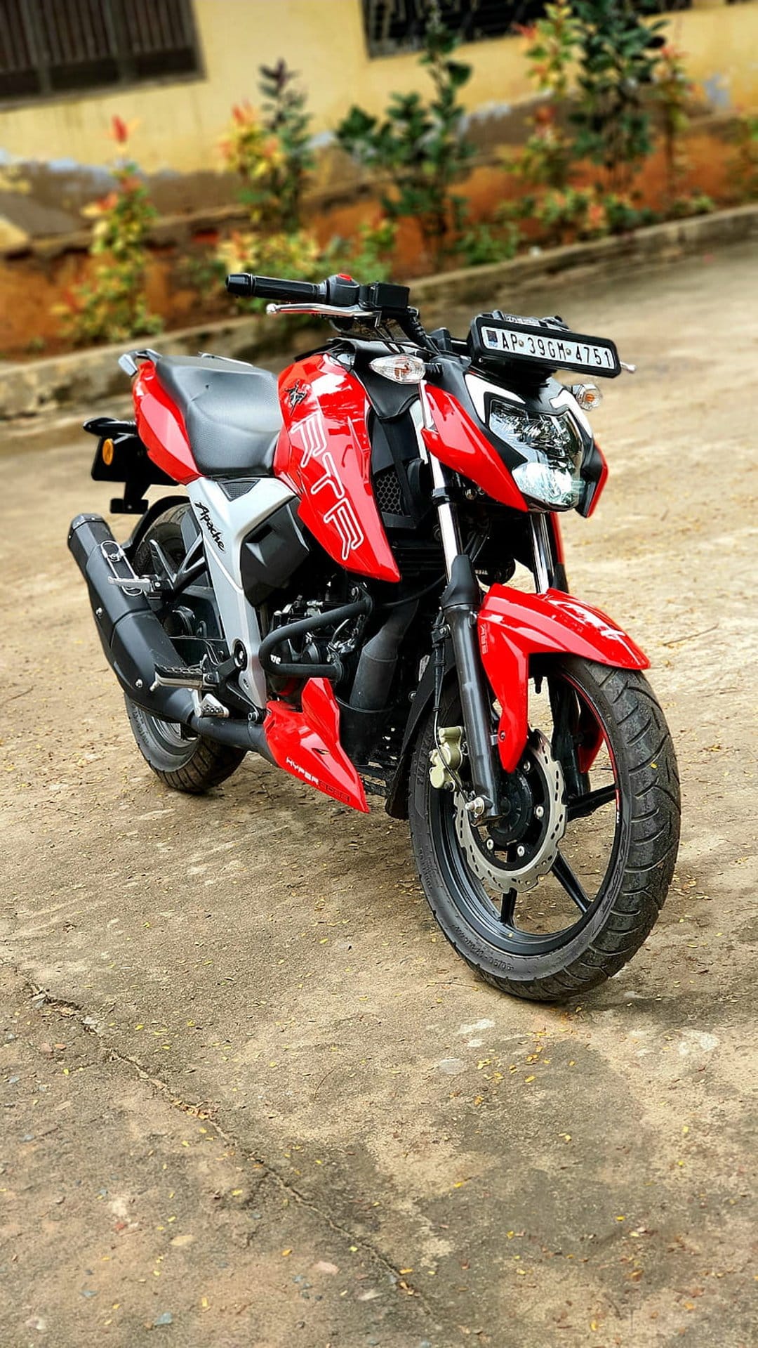Red Apache RTR motorcycle parked on cracked concrete with a blurred background of green plants and a building.