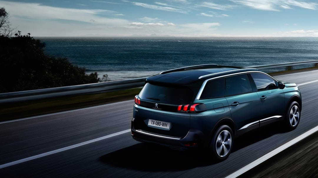 Teal SUV driving on a coastal road with motion blurred wheels, ocean waves, and a cloudy blue sky background.