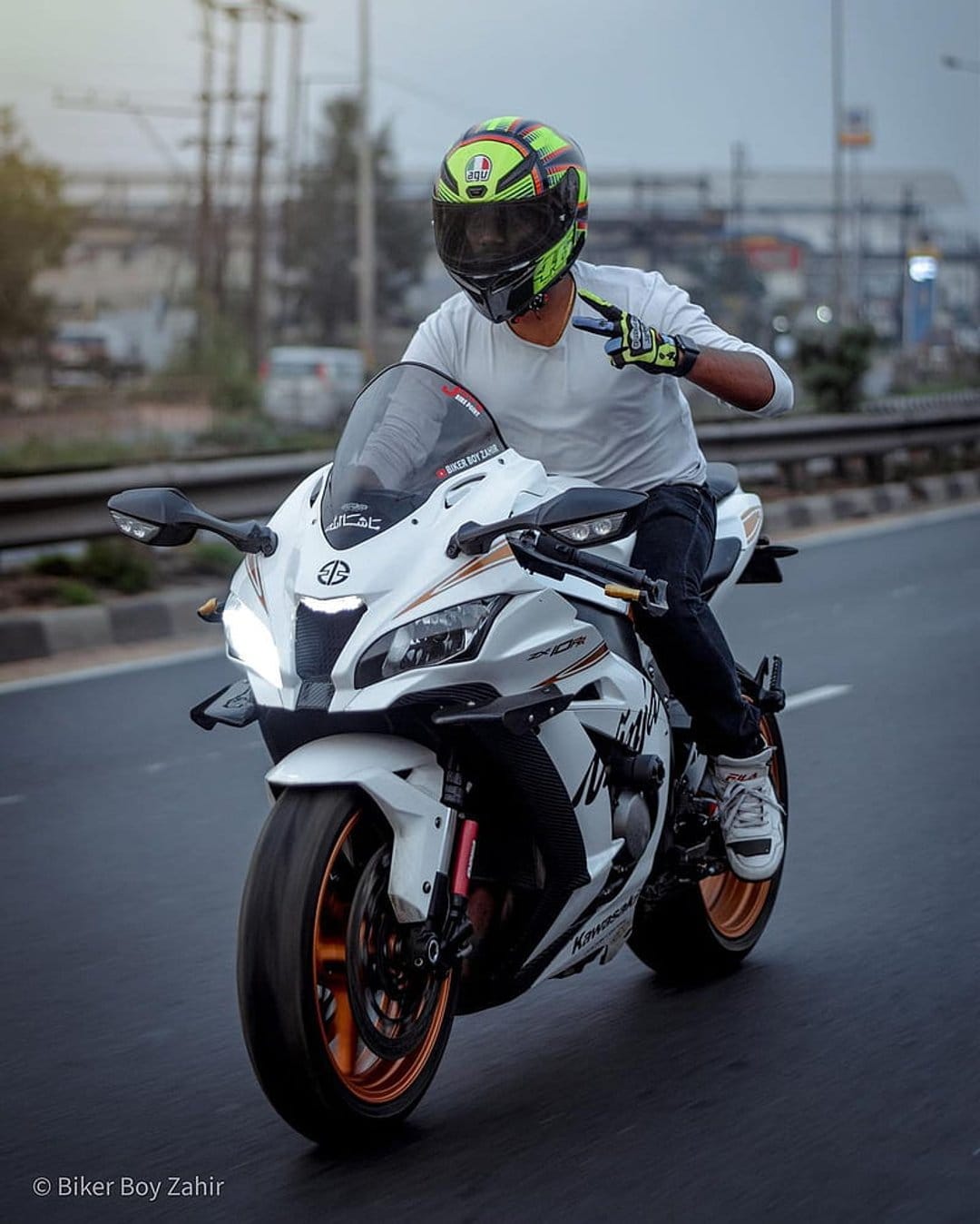 Motorcyclist in a green helmet flashing a peace sign while riding a white sportbike on a blurred city street.