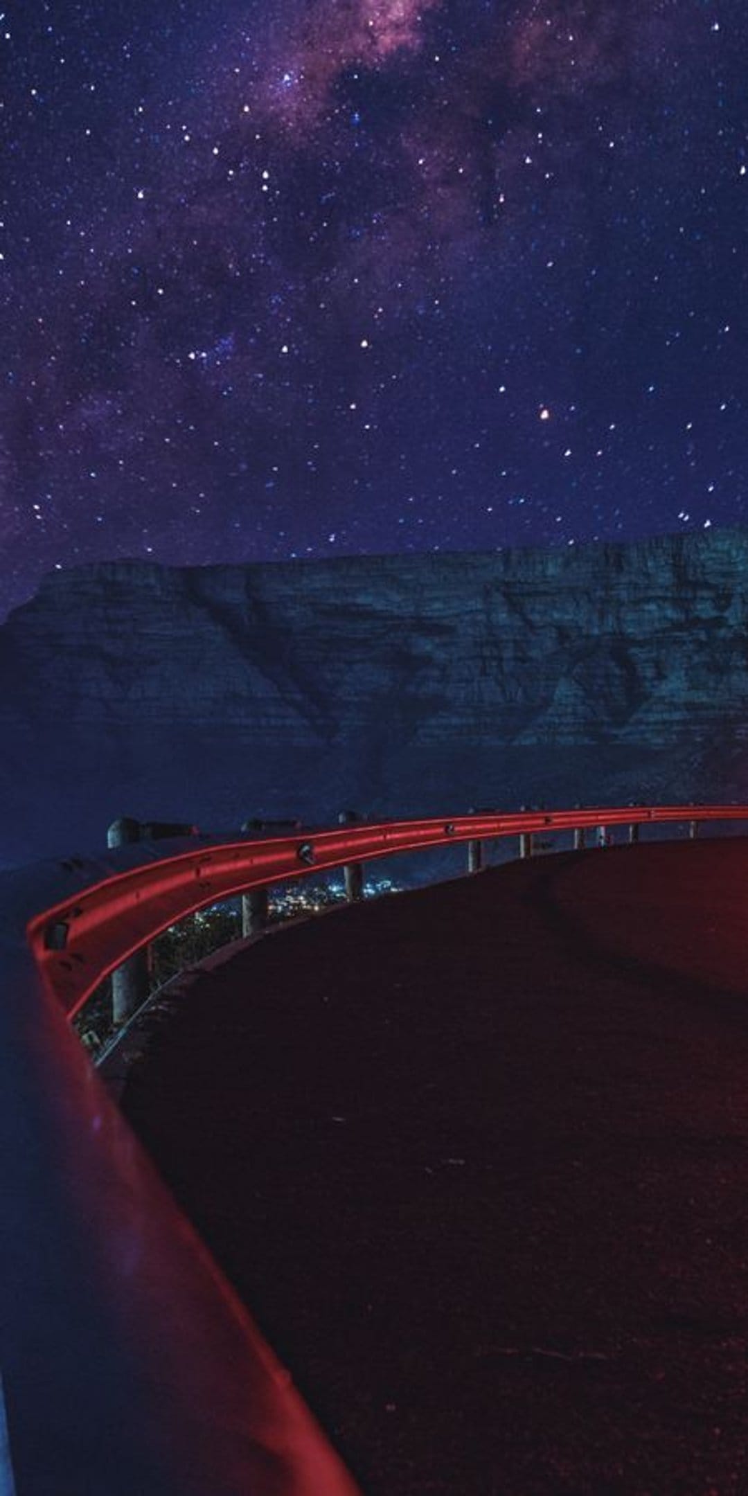Red guardrail curves along a dark asphalt road under a purple Milky Way galaxy filled with glowing star clusters.