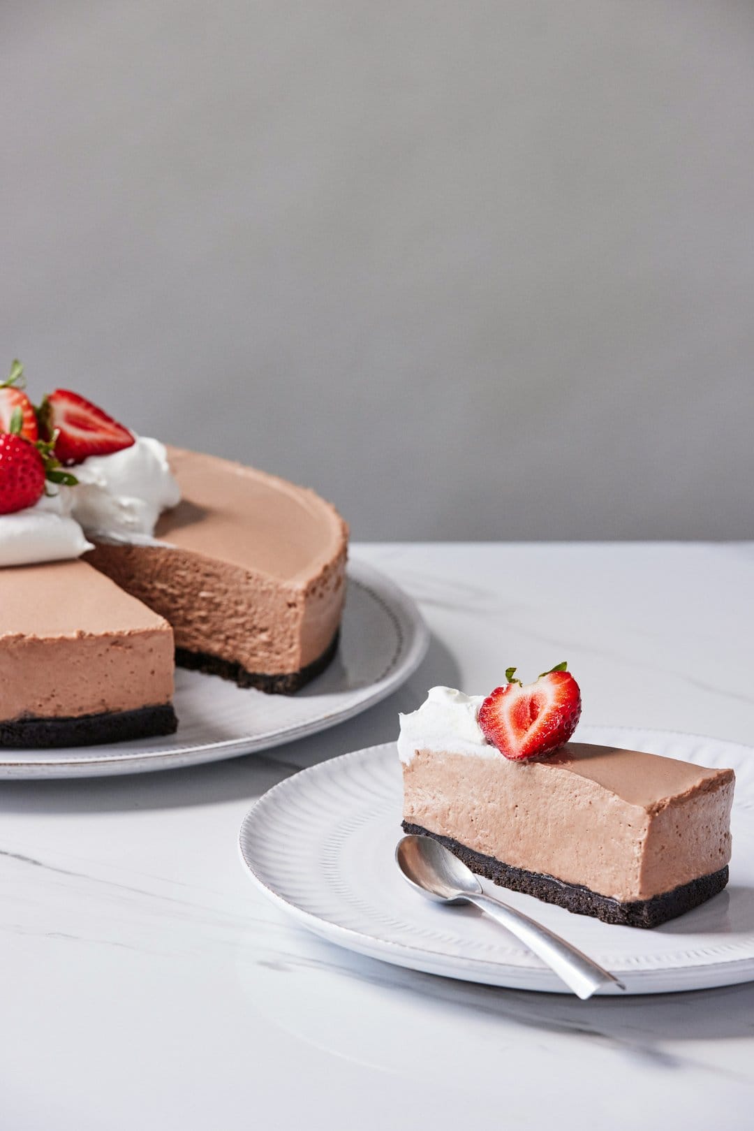 Chocolate cheesecake slice topped with whipped cream and strawberries on a white plate with a silver spoon nearby.