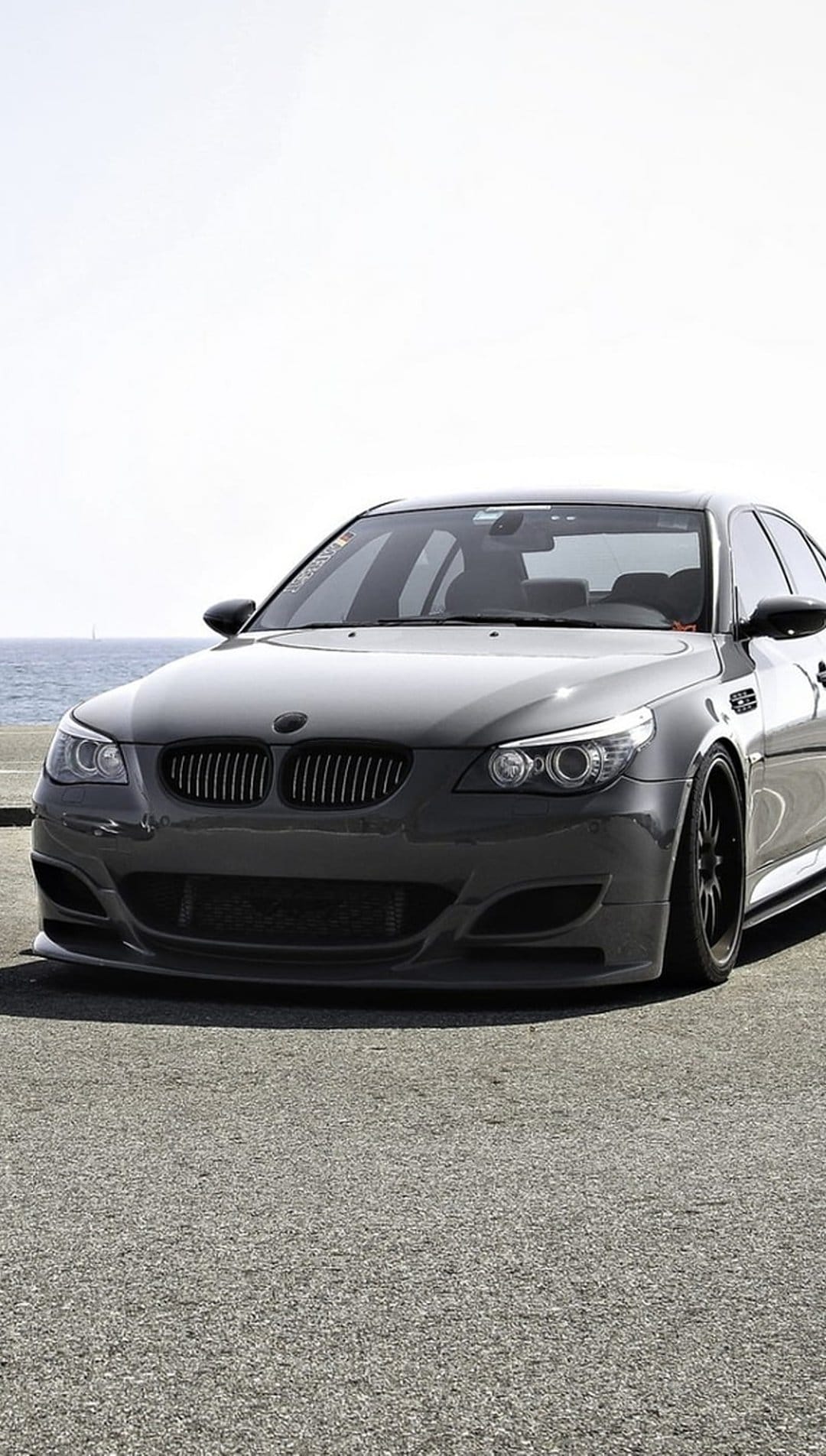 BMW M5 E60 sedan in dark gray parked on asphalt pavement beside a calm blue ocean under a clear bright sky.