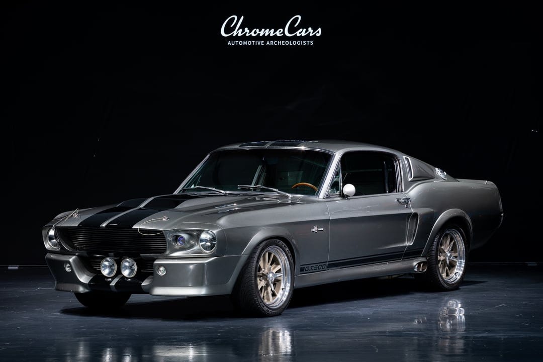 Metallic grey 1967 Shelby GT500 Eleanor with black stripes and fog lights on a dark reflective studio floor.
