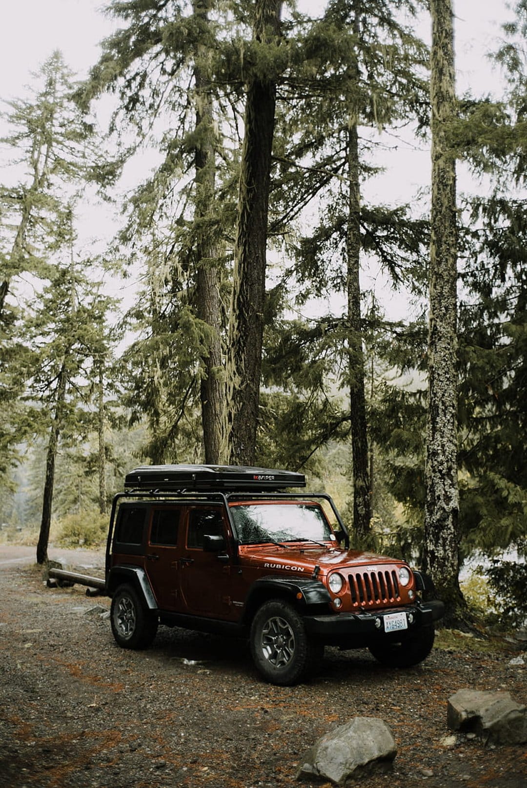Red Jeep Rubicon with a rooftop tent parked on a gravel forest road surrounded by misty moss-covered evergreen trees.